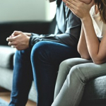 Zwei Menschen sitzen auf einem Sofa,  beide stützen ihre Arme auf den Oberschenkeln ab. Eine Person lehnt sich dabei nach vorne und hat die Hände vor dem Mund verschränkt, die Person daneben hat die Hände ineinandergelegt