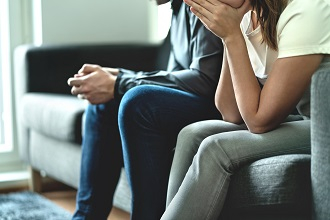 Zwei Menschen sitzen auf einem Sofa,  beide stützen ihre Arme auf den Oberschenkeln ab. Eine Person lehnt sich dabei nach vorne und hat die Hände vor dem Mund verschränkt, die Person daneben hat die Hände ineinandergelegt
